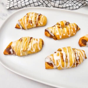 Five pumpkin crescent rolls with a cinnamon filling, drizzled with white icing, are arranged on a white plate. A checkered kitchen towel is in the background.