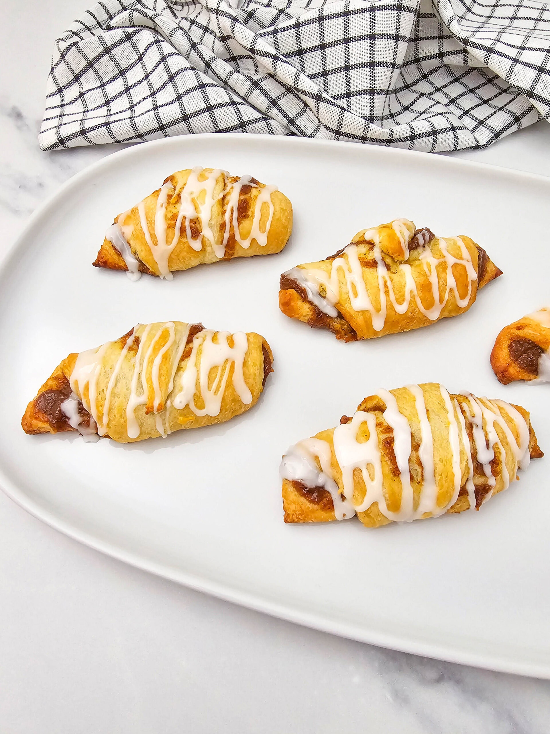Five pumpkin crescent rolls with a cinnamon filling, drizzled with white icing, are arranged on a white plate. A checkered kitchen towel is in the background.