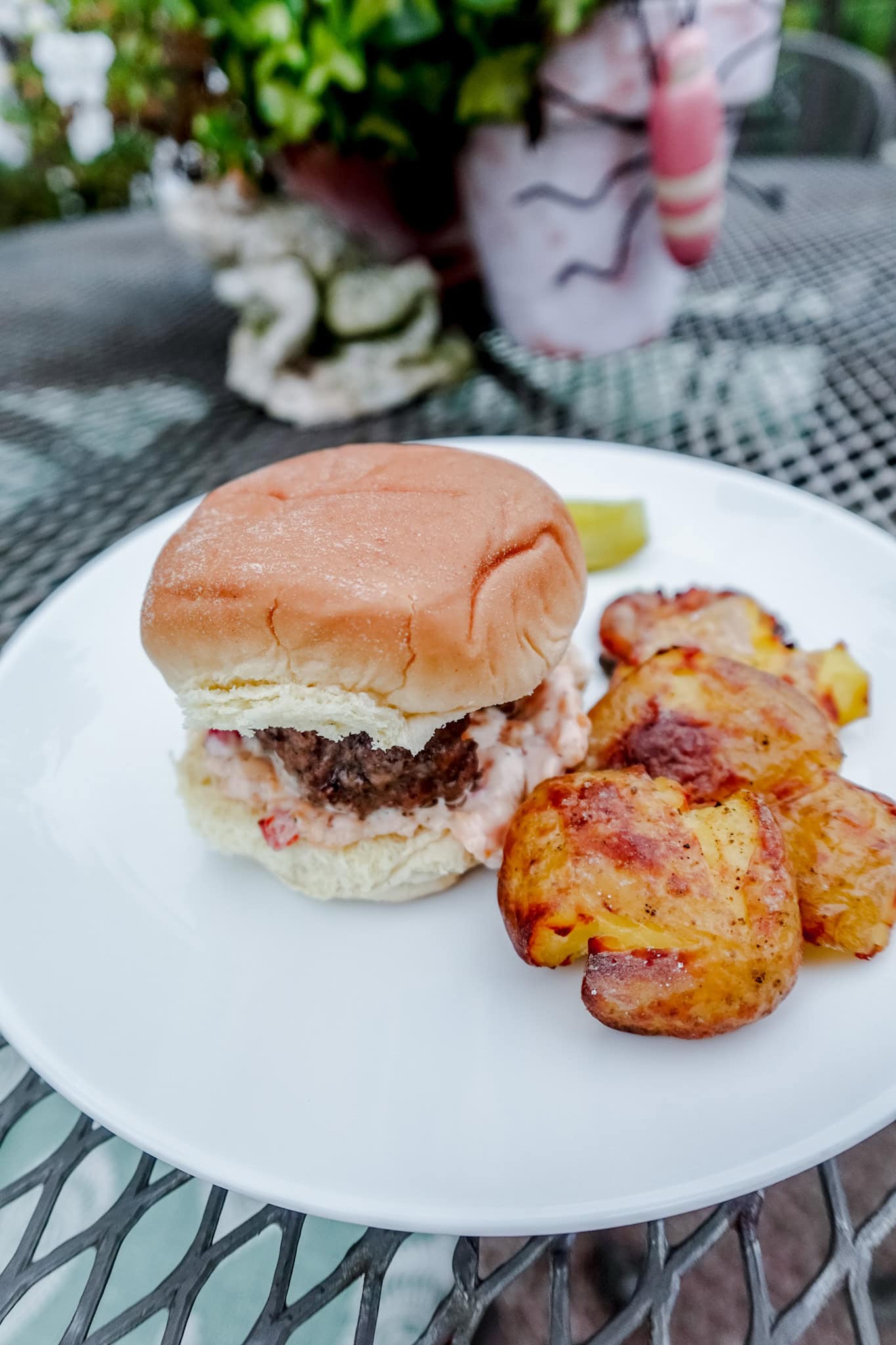 A juicy burger with a creamy sauce sits on a plate beside golden-brown roasted potatoes, garnished with a pickle slice.