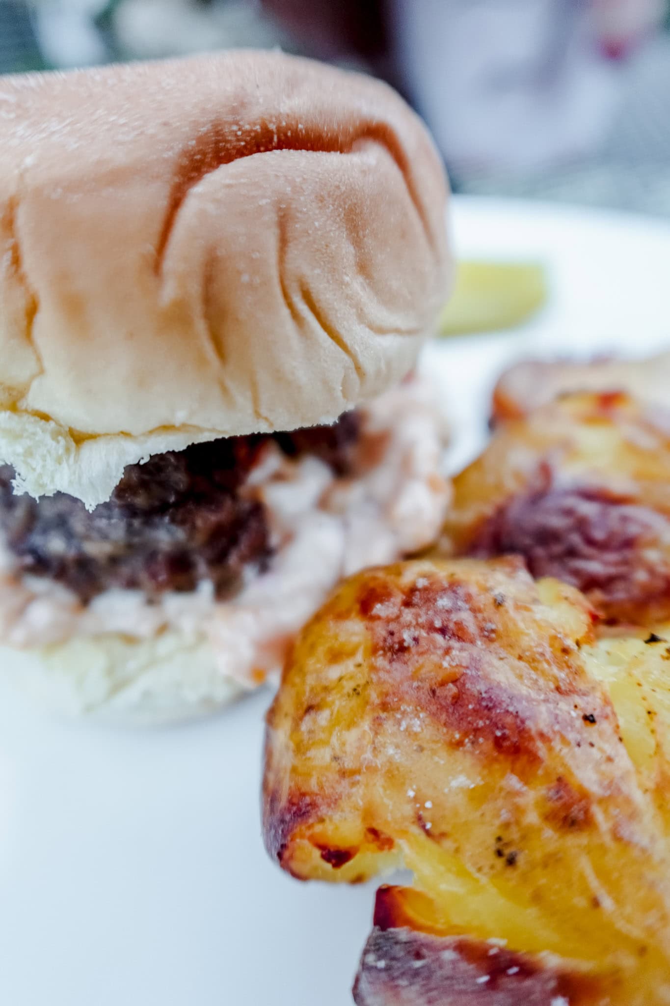 A close-up of a soft hamburger bun with a savory patty and sauce, accompanied by golden-brown roasted potatoes on a white plate.