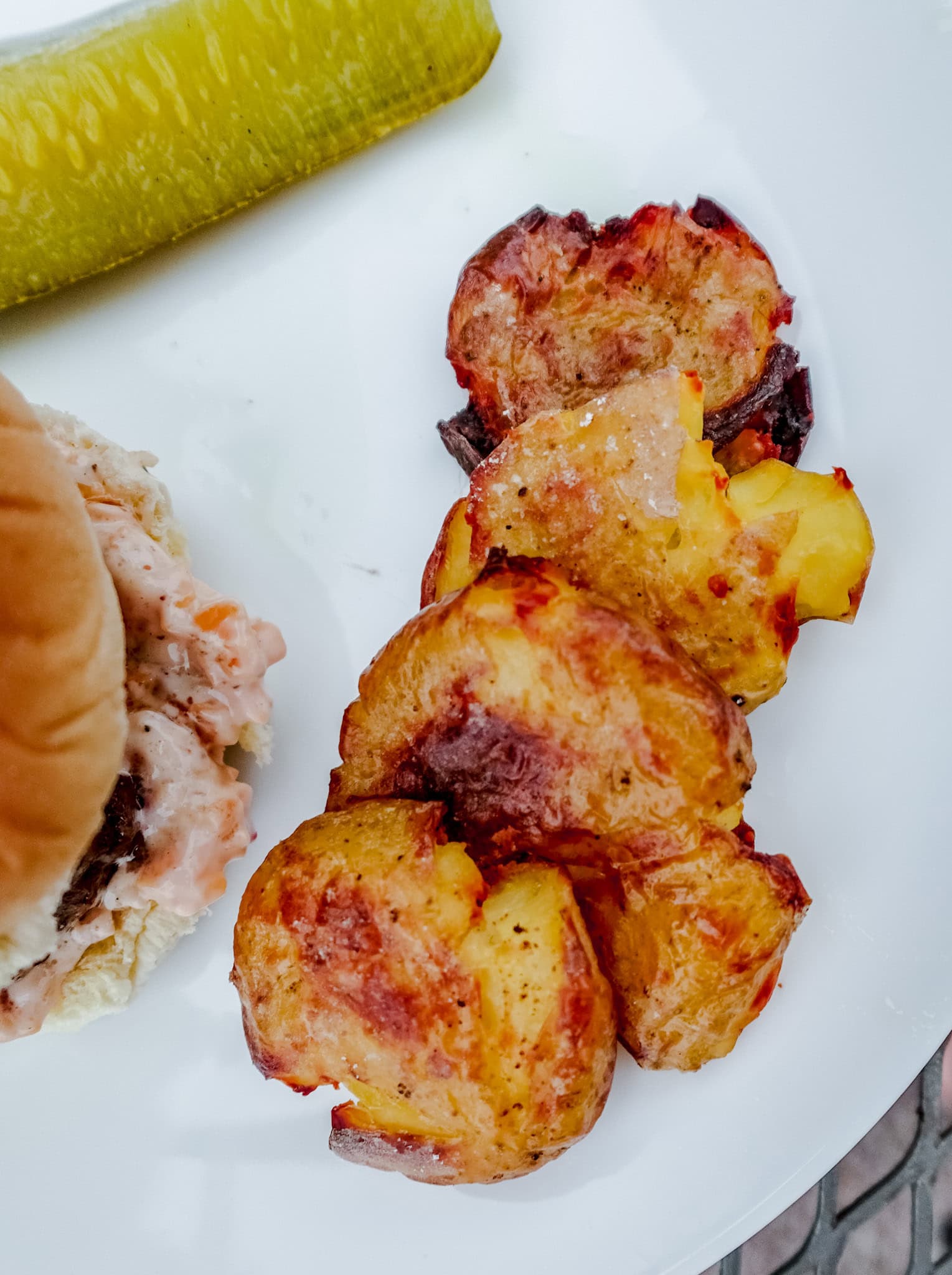 A plate featuring a burger with creamy dressing, crispy smashed potatoes, and a bright green pickle on the side.