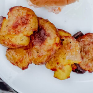 A plate with cheesy golden-brown crispy potato slices, garnished and ready to be served, alongside a sandwich and pickle.