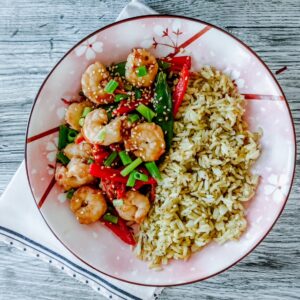 A floral plate with shrimp stir-fry, red peppers, green onions, and snow peas on the left, and seasoned brown rice on the right atop a white and blue napkin set on a gray wooden surface.