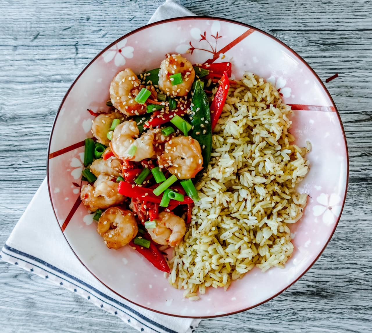 A floral plate with shrimp stir-fry, red peppers, green onions, and snow peas on the left, and seasoned brown rice on the right atop a white and blue napkin set on a gray wooden surface.
