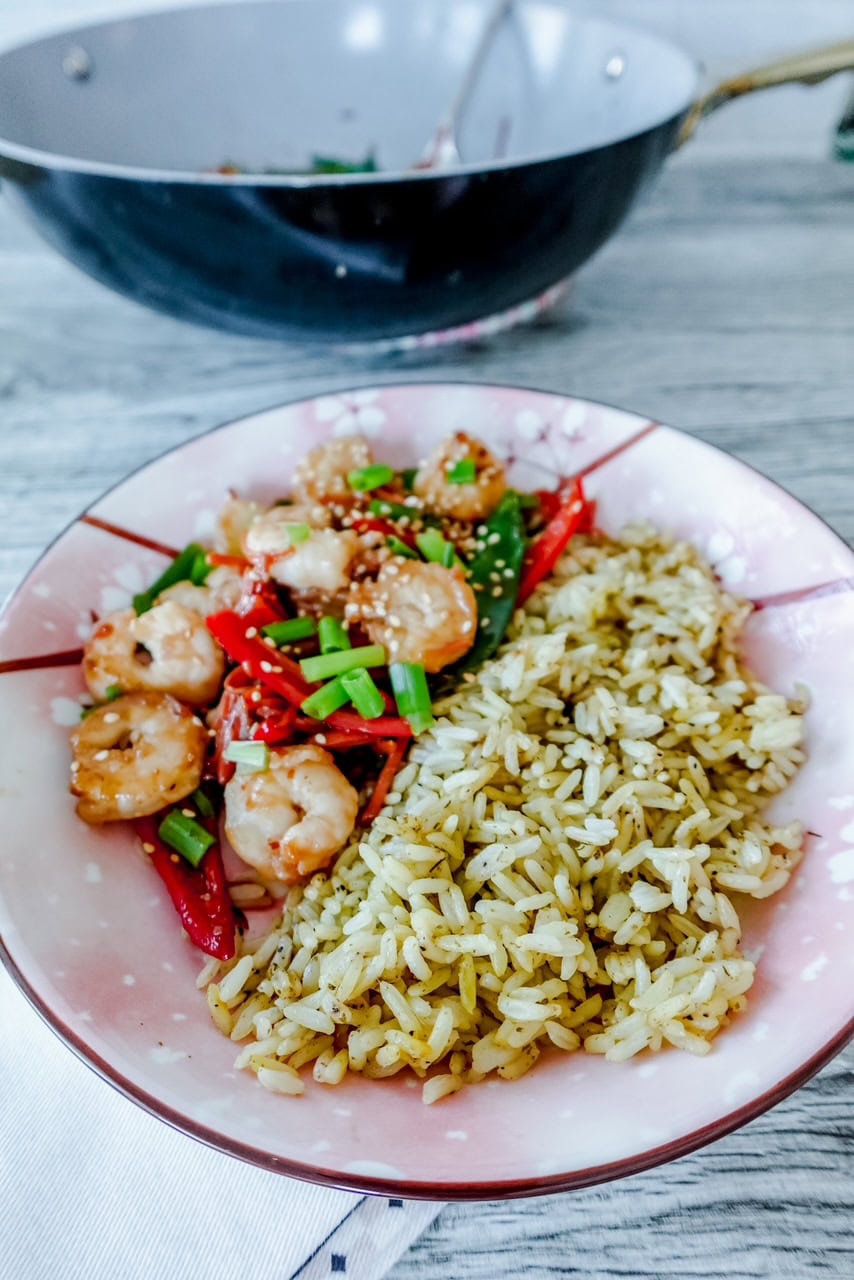A pink plate with cherry blossoms is full of shrimp stir-fry with red peppers, green onions, and sesame seeds next to a portion of seasoned rice on a wooden table. A black metal wok is in the background.