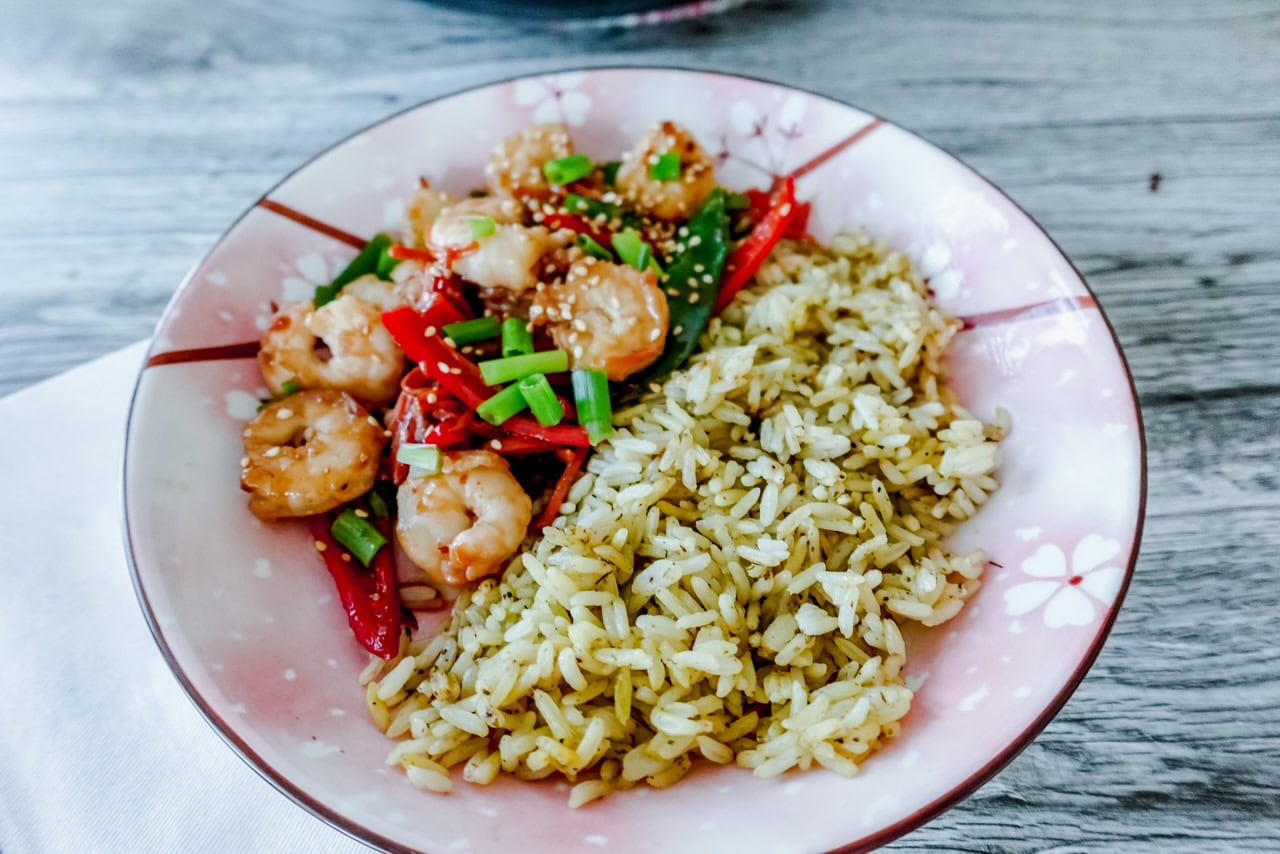 A floral-patterned plate with seasoned rice and stir-fried shrimp, red bell peppers, and green onions, garnished with sesame seeds on a wooden table.
