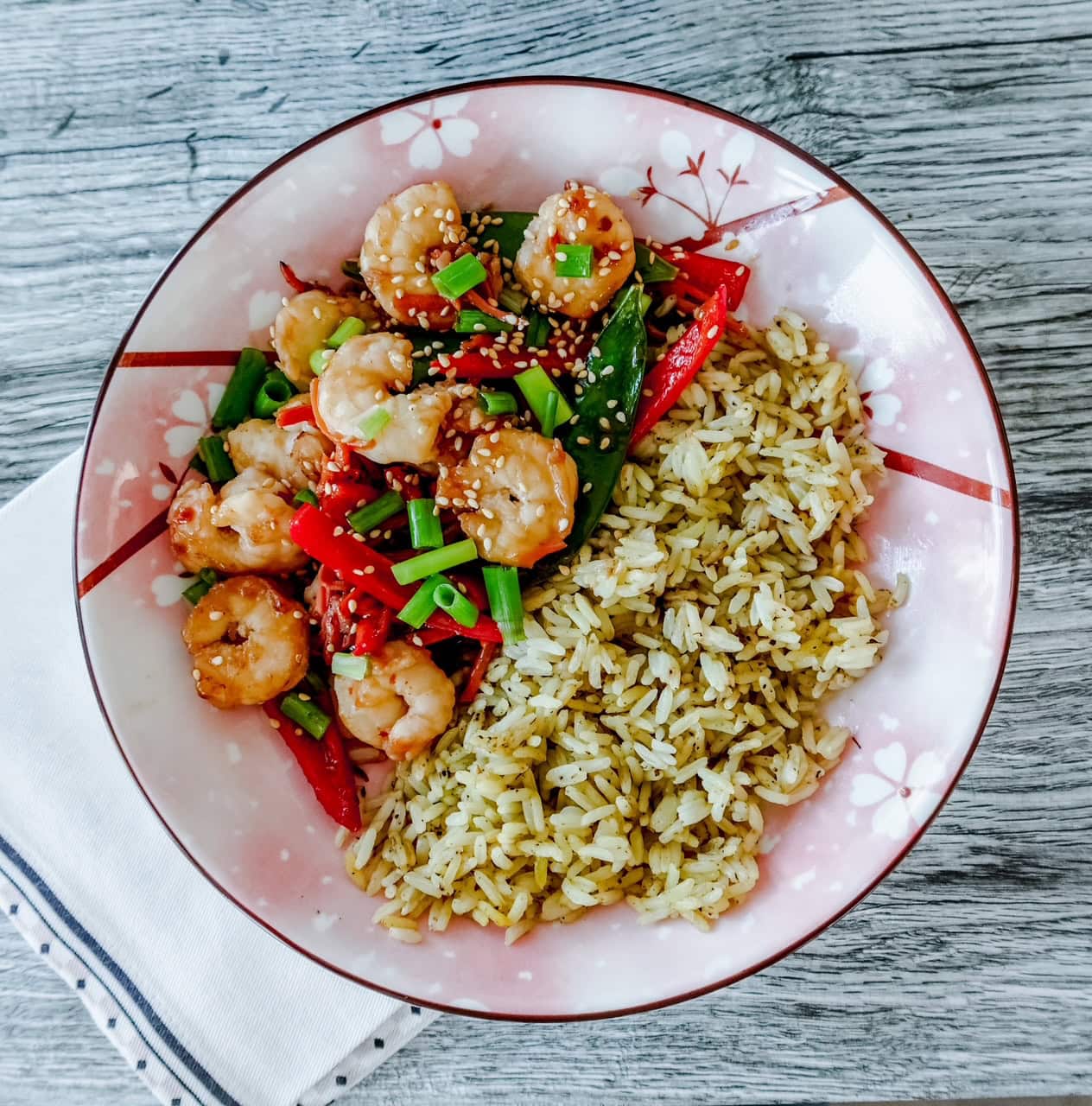 A floral-patterned plate with seasoned rice and stir-fried shrimp, red bell peppers, and green onions, garnished with sesame seeds on a white and blue napkin on a wooden table.