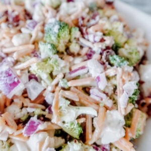 A close-up of a salad featuring chopped broccoli, shredded carrots, diced red onions, bacon bits, cheese, and a creamy dressing on a white plate.