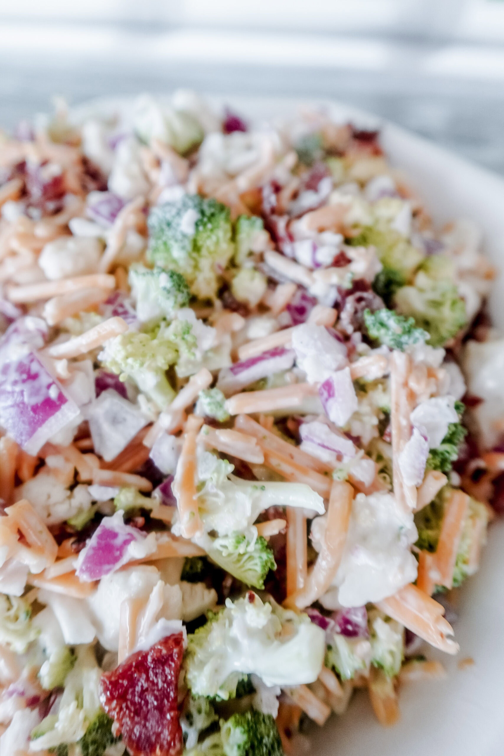 A close-up of a salad featuring chopped broccoli, shredded carrots, diced red onions, bacon bits, cheese, and a creamy dressing on a white plate.