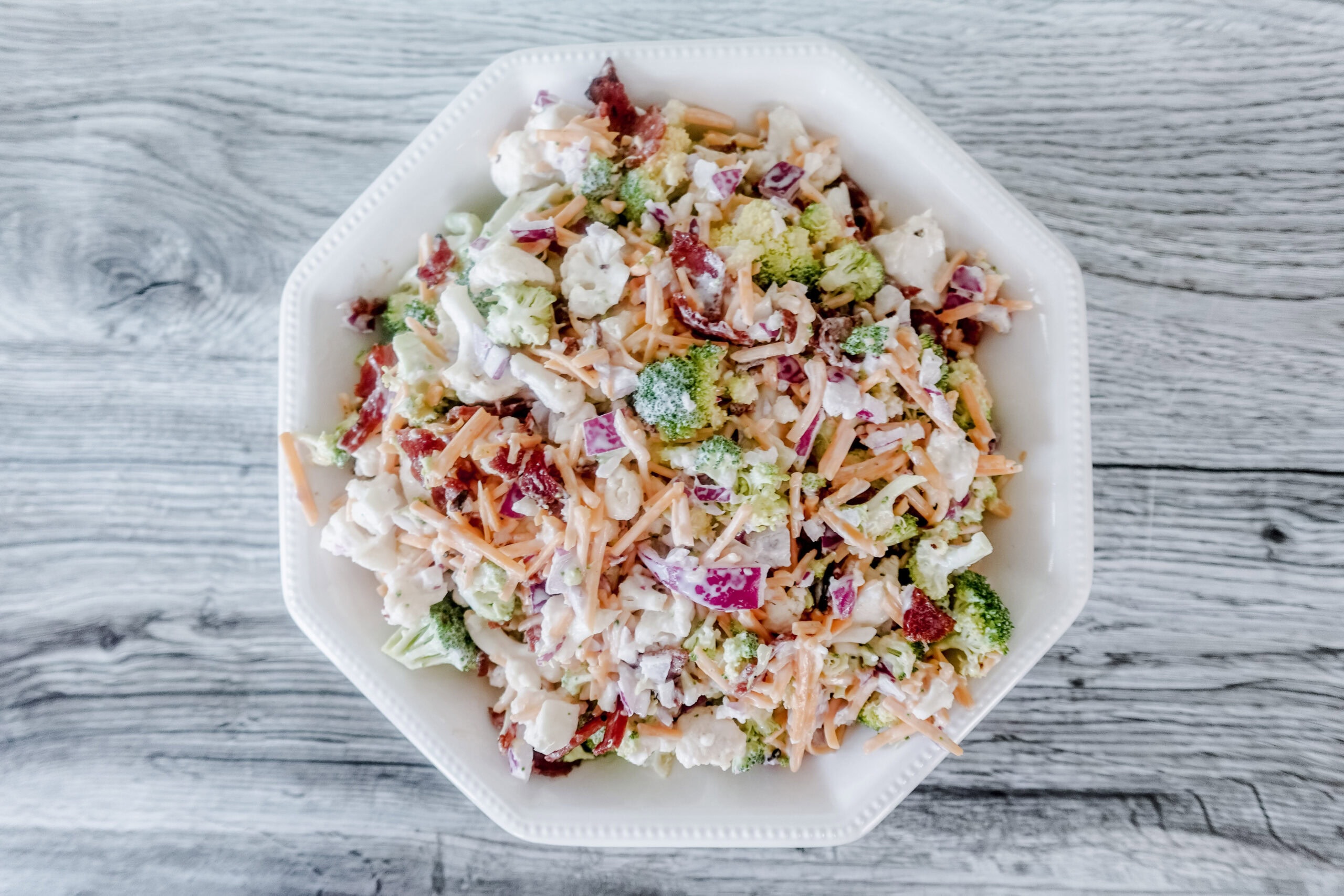 A white bowl filled with a colorful salad containing chopped broccoli, red onions, shredded carrots, bacon pieces, and a creamy dressing, set on a light wooden surface.