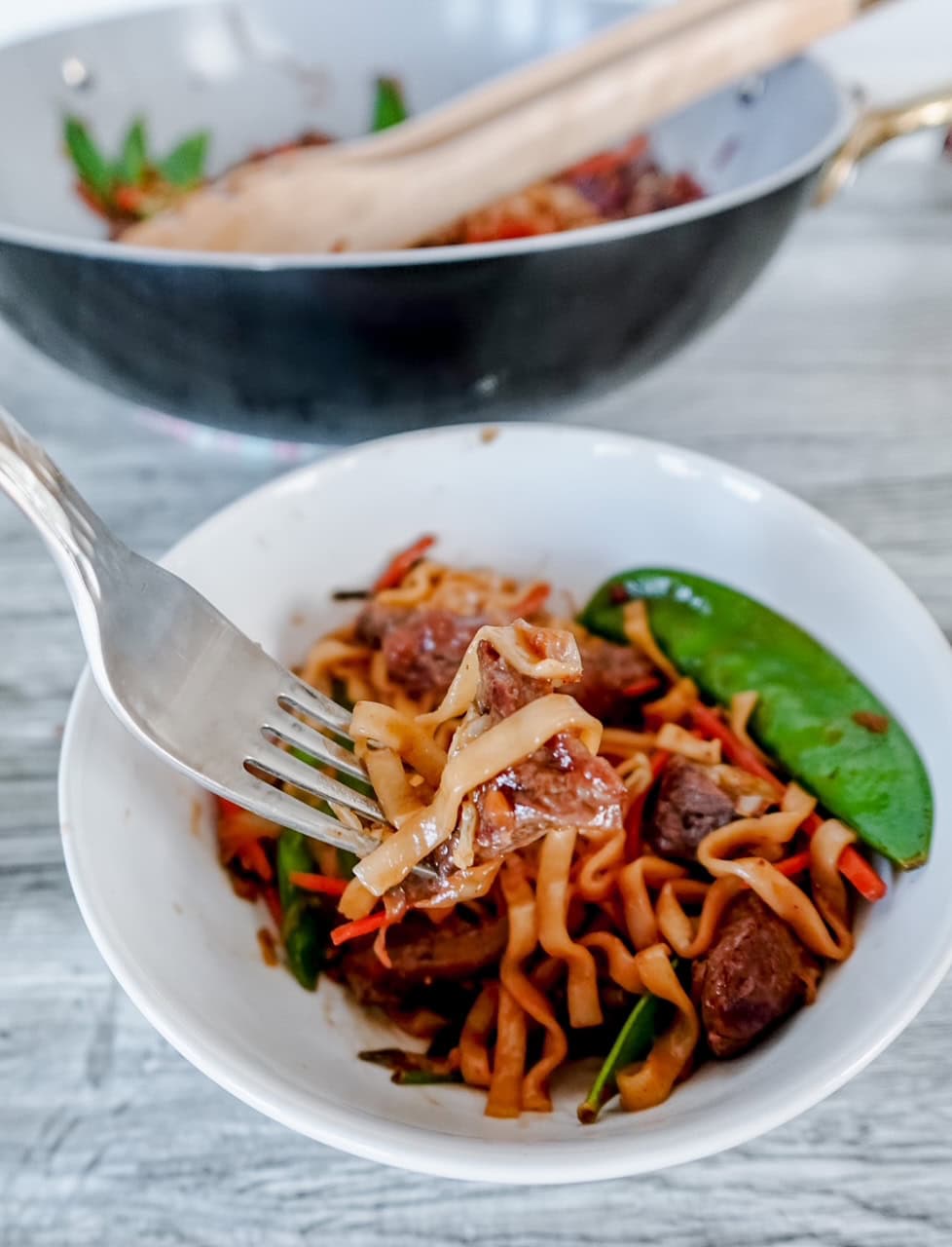 A fork holds a bite of stir-fried noodles with beef, snow peas, and vegetables above a white bowl; a wok with more of the dish sits in the background on a light wooden surface.