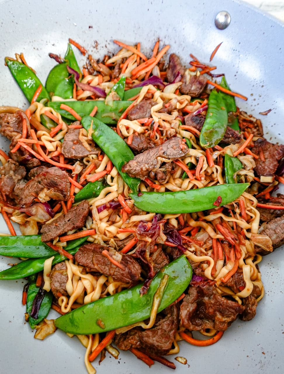 A wok filled with stir-fried beef, snow peas, shredded carrots, cabbage, and noodles in a savory sauce.