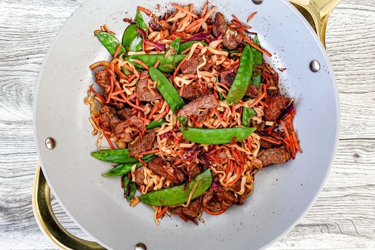 A white wok filled with stir-fried noodles, beef strips, snap peas, shredded carrots, and cabbage, placed on a light wooden surface.