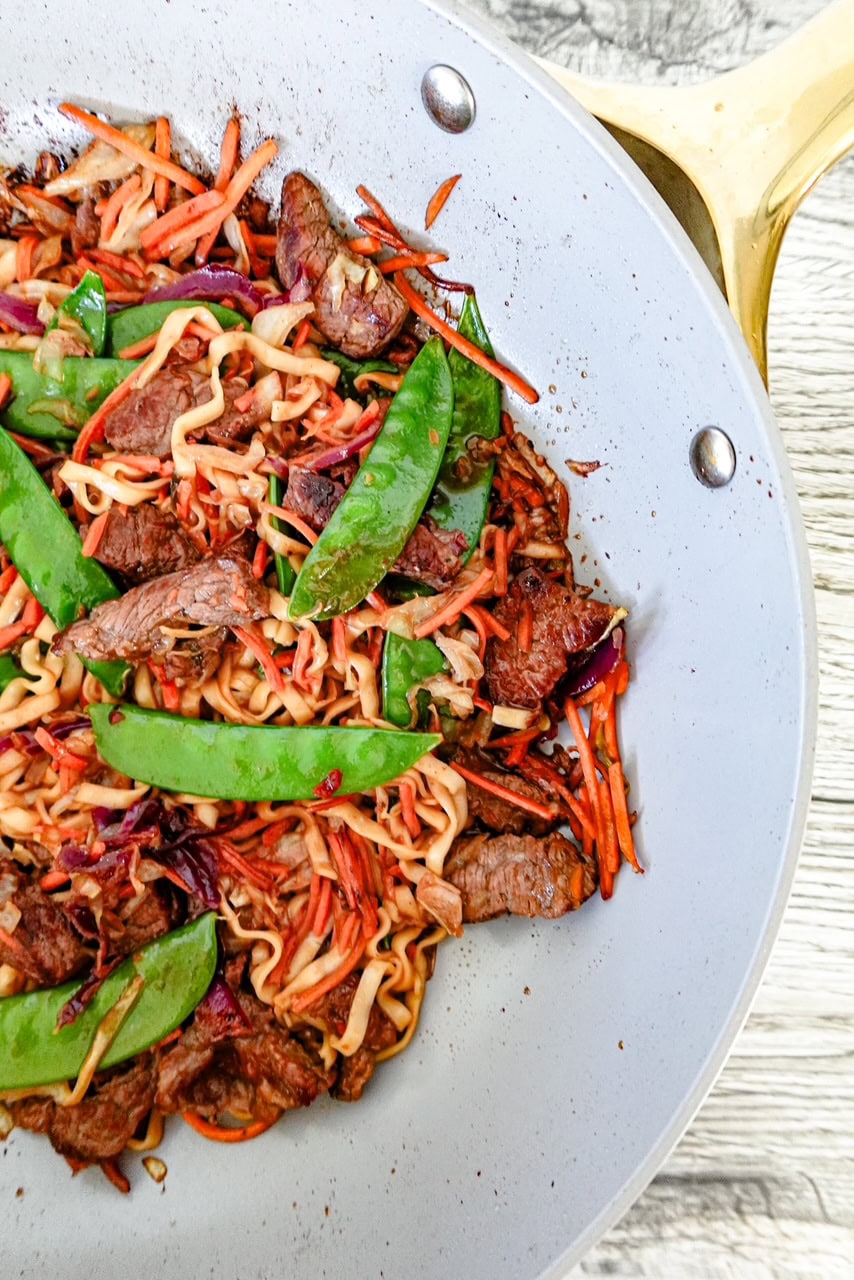 A close-up of a stir-fry in a white skillet with gold handle, featuring sliced beef, ramen noodles, snap peas, shredded carrots, red cabbage, and greens on a light wood surface.