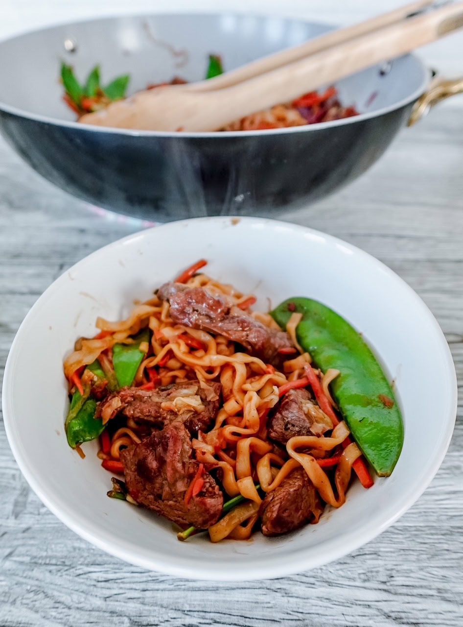 A bowl of stir-fried noodles with beef, snap peas, and vegetables sits on a light surface, with a wok and wooden tongs in the background.