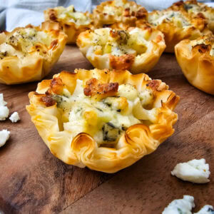 Mini phyllo pastry cups filled with melted cheese and pears are arranged on a wooden board, with a few pieces of crumbled blue cheese scattered nearby.