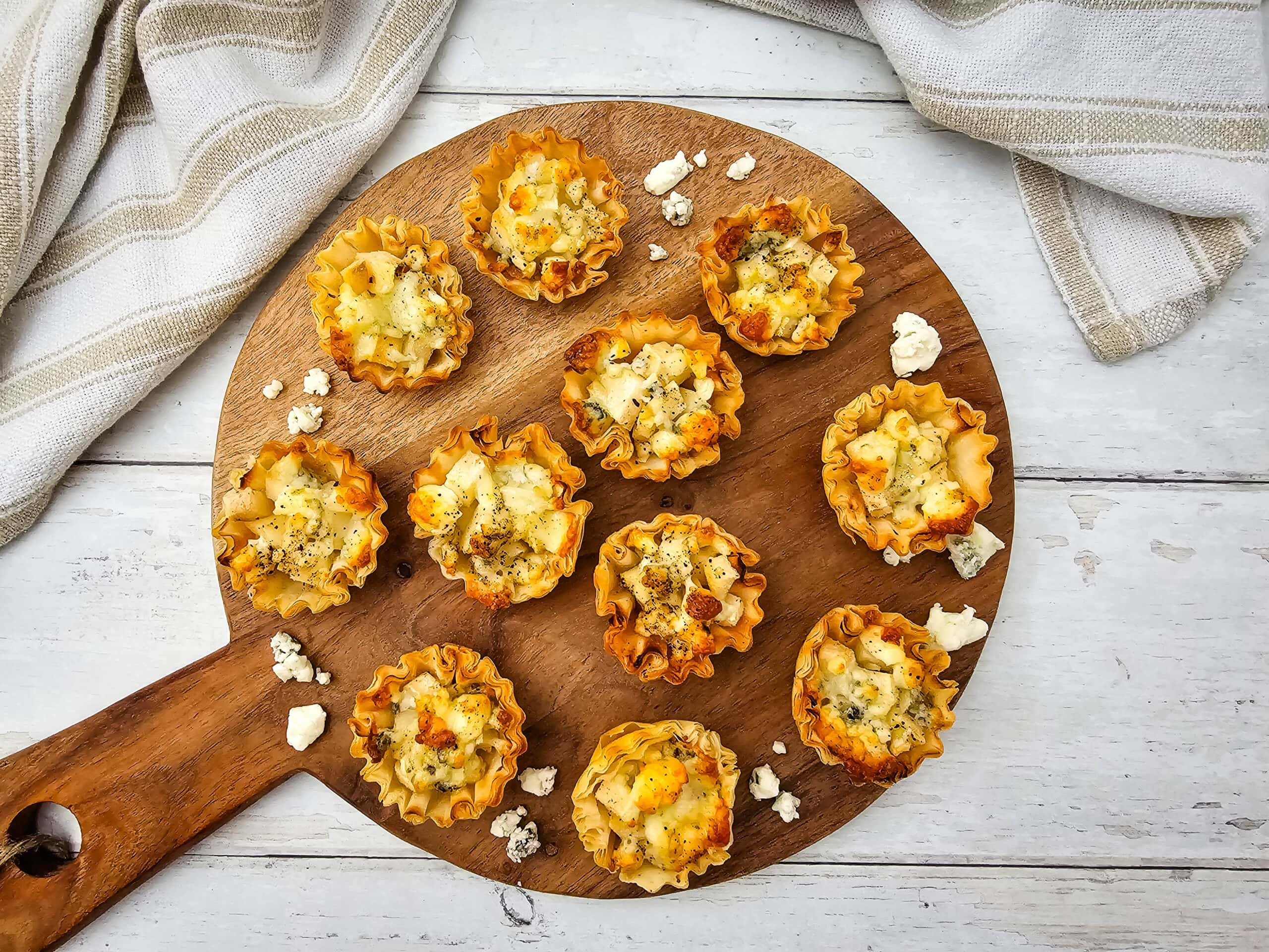 An overhead shot of mini phyllo pastry cups filled with cheese and pears are arranged on a round wooden serving board, with a few blue cheese crumbles scattered around. The board sits on a white wooden surface with striped towels nearby.