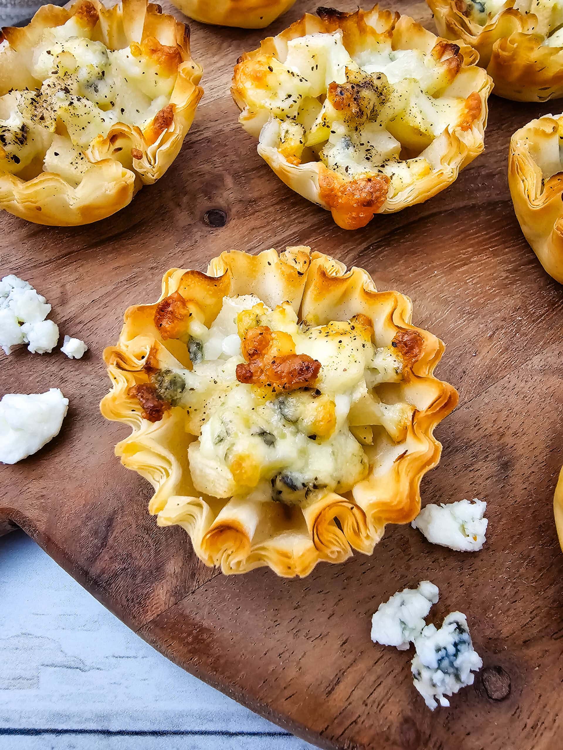 An overhead shot of mini phyllo pastry cups filled with a cheesy mixture and diced pears, baked to a golden brown, are arranged on a wooden board. Some crumbled blue cheese is scattered around the cups.