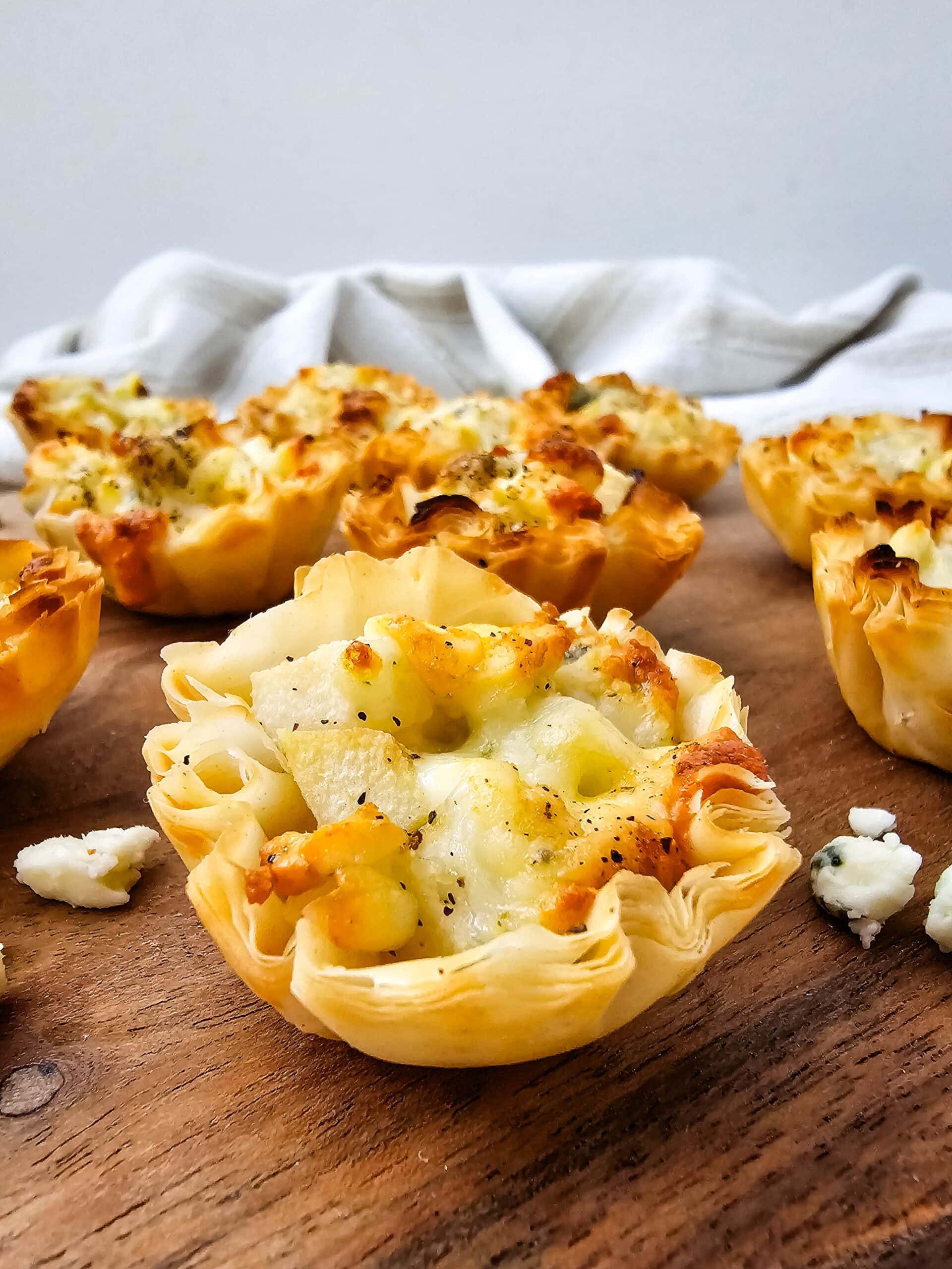 Mini phyllo pastry cups filled with melted blue cheese and diced pears are arranged on a wooden surface with a light cloth in the background. The pastries are golden and slightly crisp around the edges.