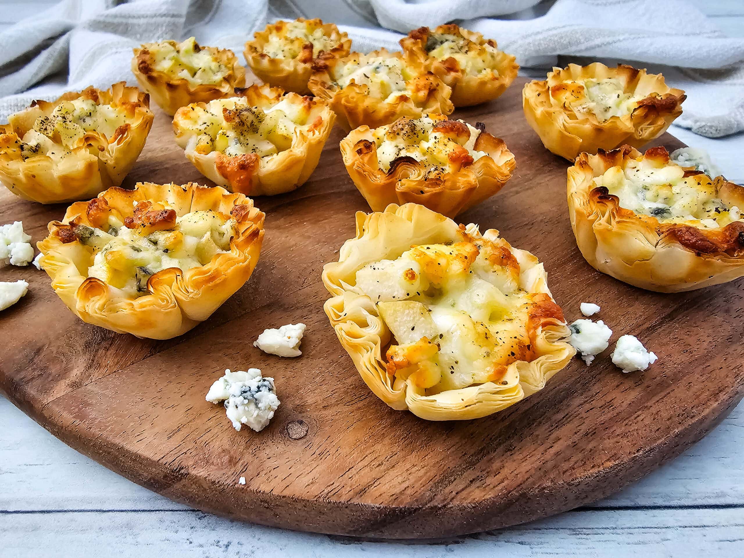 Eight baked phyllo pastry cups filled with blue cheese and pears sit on a round wooden board, with small crumbles of blue cheese scattered around, against a light wooden background.