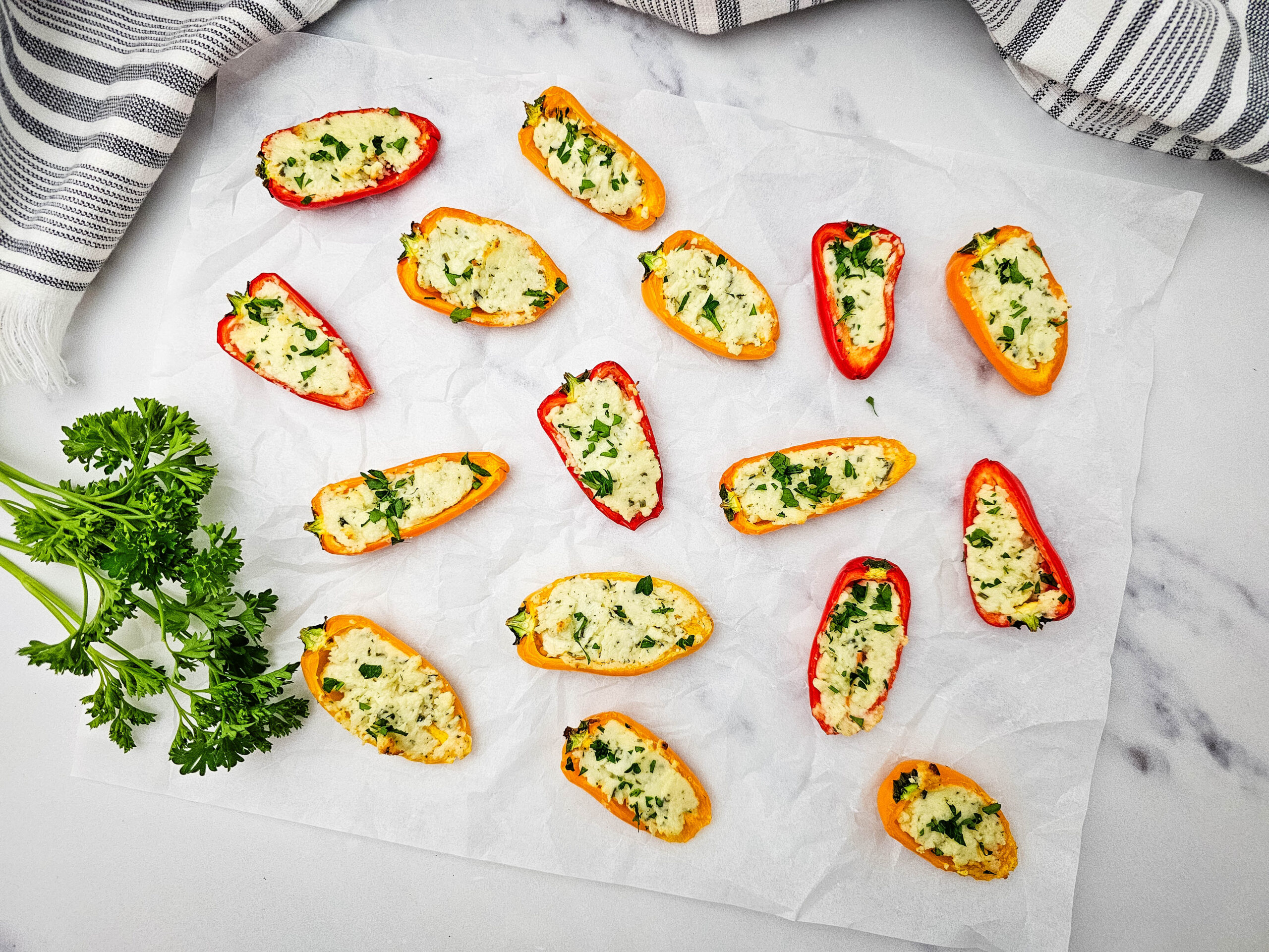 An overhead shot of stuffed mini bell peppers filled with a creamy cheese and herb mixture are arranged on parchment paper. A bunch of fresh parsley sits nearby, and a striped kitchen towel is in the background.
