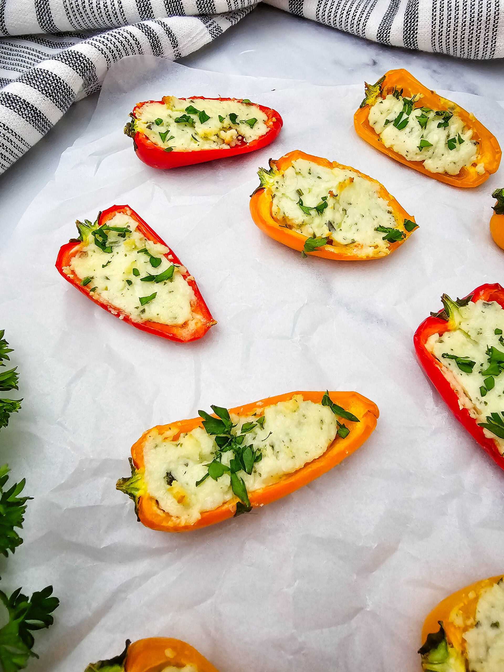 Colorful mini bell peppers halved and filled with a creamy cheese mixture, garnished with chopped herbs, are arranged on parchment paper with a striped cloth and fresh parsley nearby.