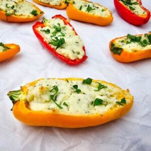 Colorful halved mini bell peppers filled with a creamy, herbed cheese mixture and topped with chopped parsley, arranged on a sheet of white parchment paper.