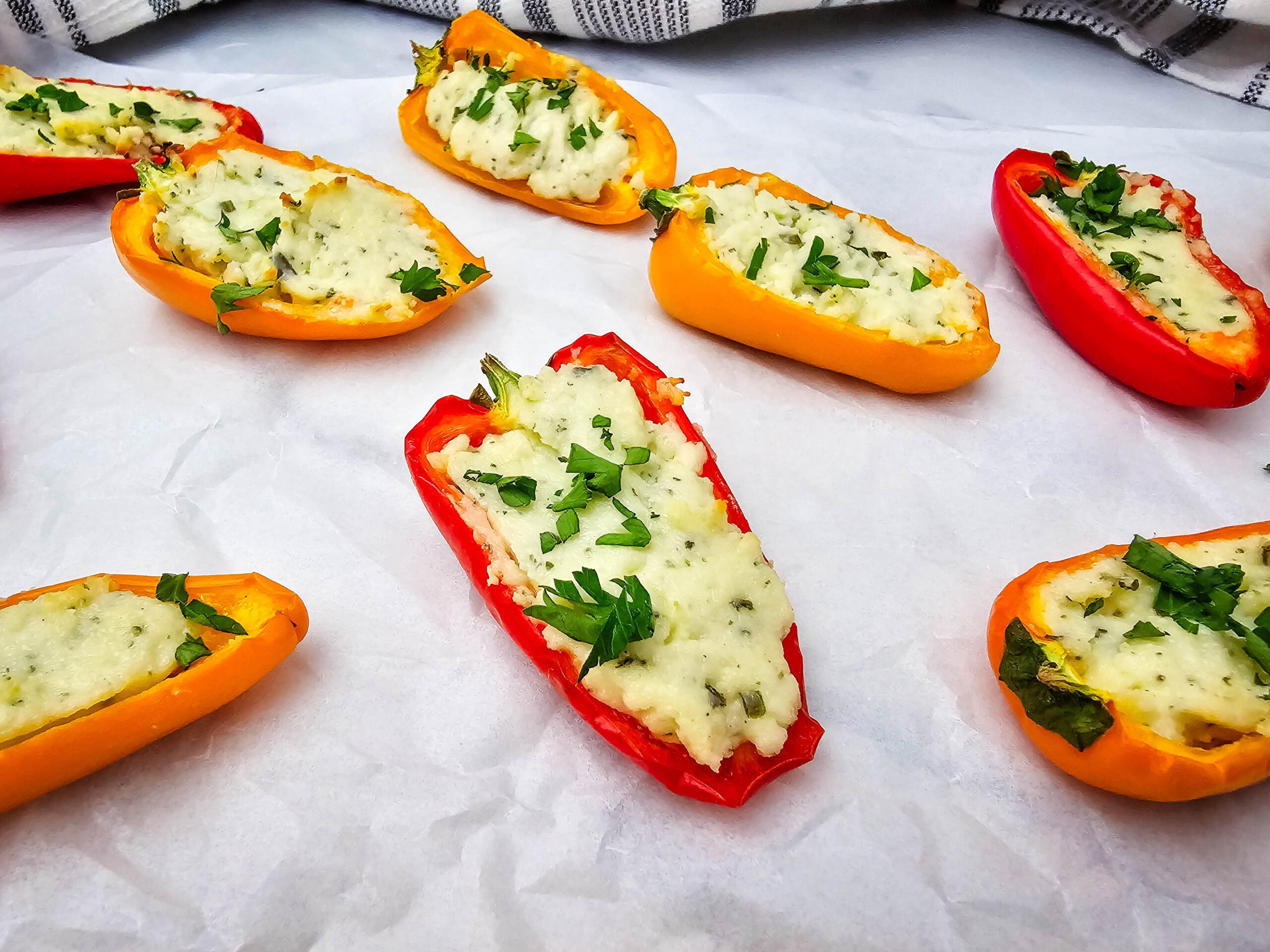 Colorful mini bell peppers halved and filled with a cheesy, herb-topped stuffing, arranged on white parchment paper with a striped cloth in the background.