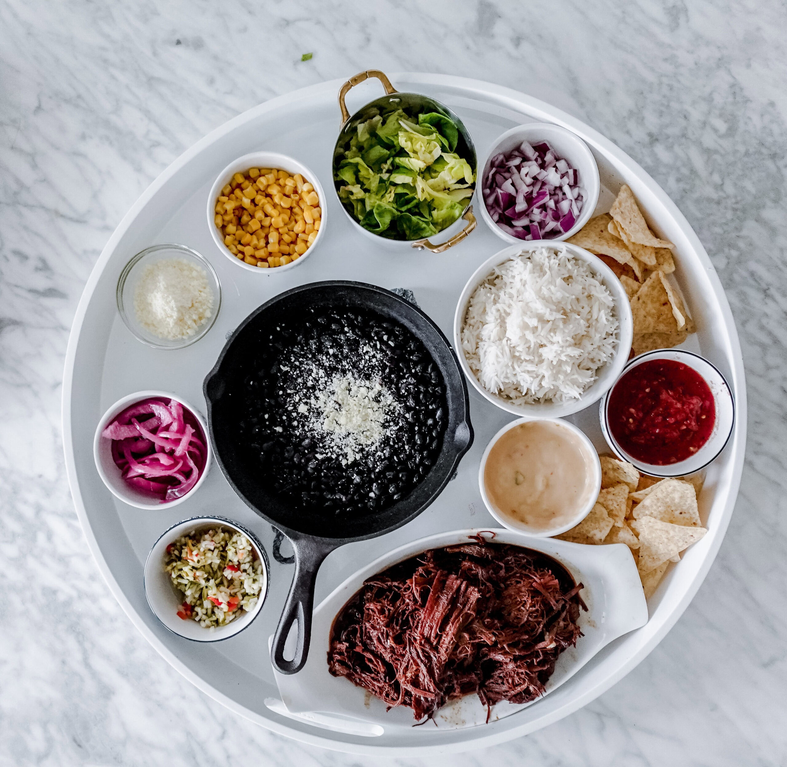 An overhead shot of a round tray with bowls of shredded beef, black beans, rice, corn, lettuce, chopped onions, salsa, pickled onions, chips, sauces, grated cheese, and a vegetable mix arranged neatly on a marble surface.