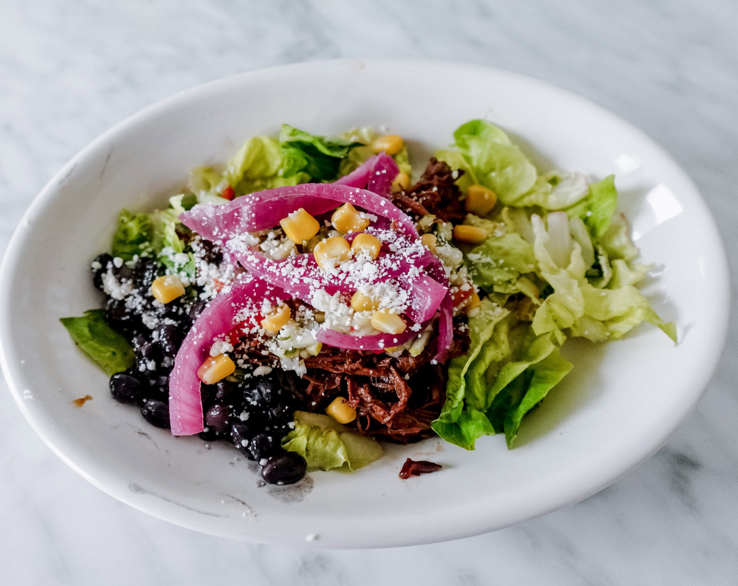 A close-up of a white bowl with lettuce, shredded beef, black beans, pickled red onions, corn, and crumbled cheese, served on a marble surface.