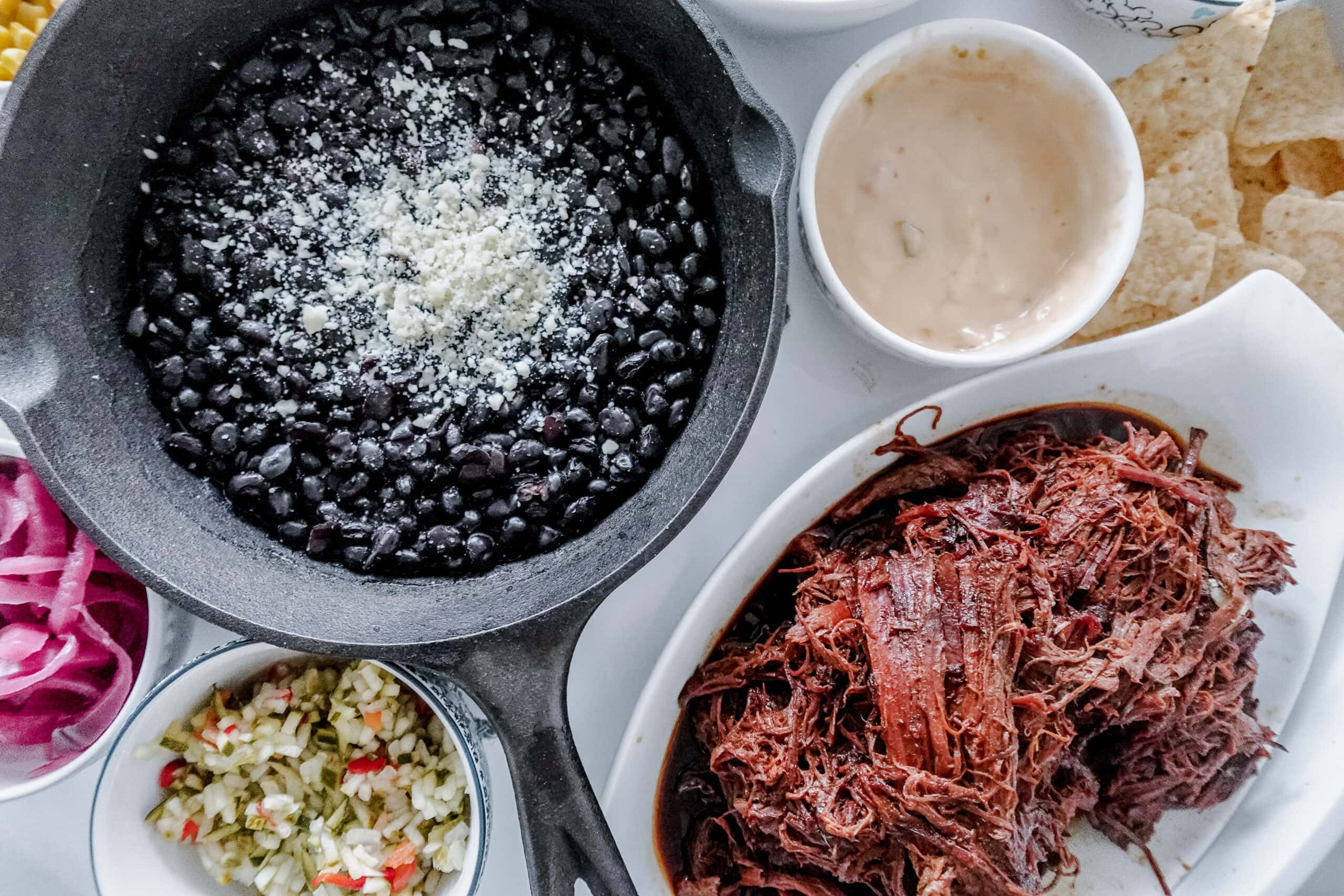A close-up of a meal with a skillet of black beans topped with cheese, a bowl of shredded beef, pickled onions, chopped vegetables, a cheese dipping sauce, and tortilla chips on a white serving dish.
