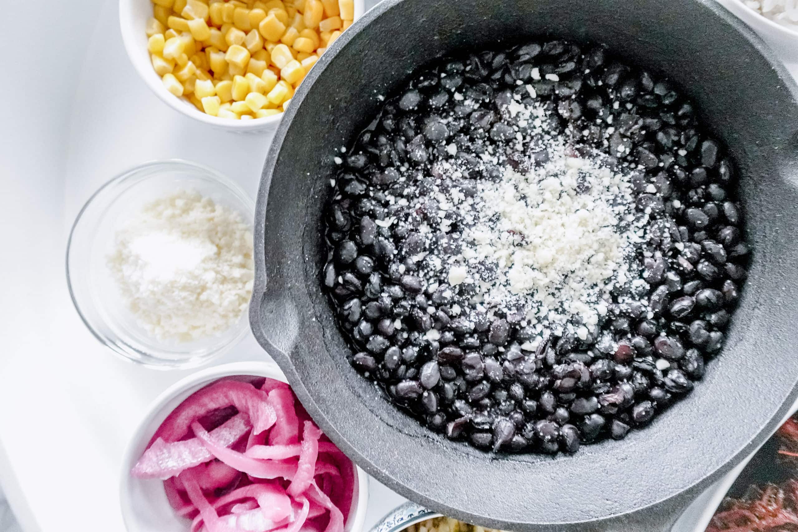 A black skillet filled with black beans topped with grated cheese, surrounded by small bowls of corn, pickled red onions, and grated cheese on a white serving dish.