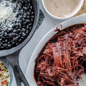 A close-up of a meal featuring shredded beef in a white dish, black beans with cheese in a cast iron skillet, cheesy dip with tortilla chips, and a small bowl of vegetables.