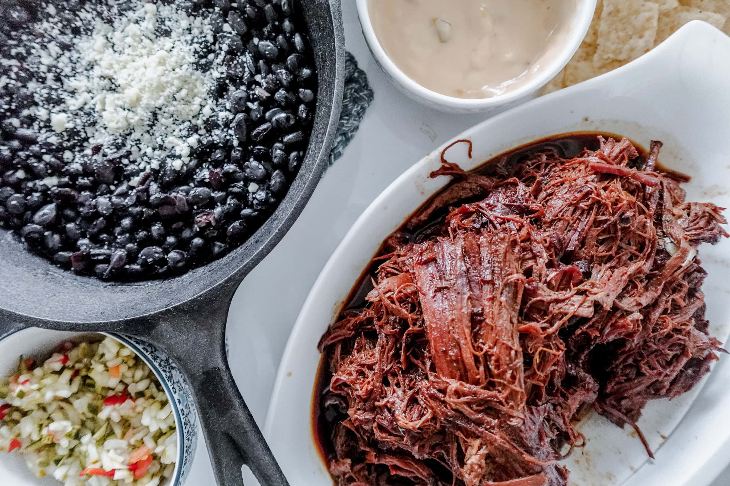 A close-up of a meal featuring shredded beef in a white dish, black beans with cheese in a cast iron skillet, cheesy dip with tortilla chips, and a small bowl of vegetables.