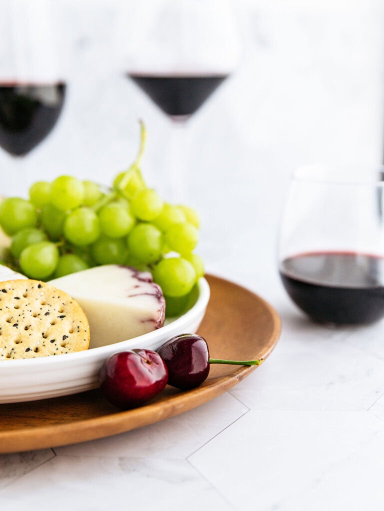 A plate with green grapes, cheese, crackers, and cherries sits on a wooden tray. In the background, three glasses of red wine are partially visible against a light, blurred backdrop.