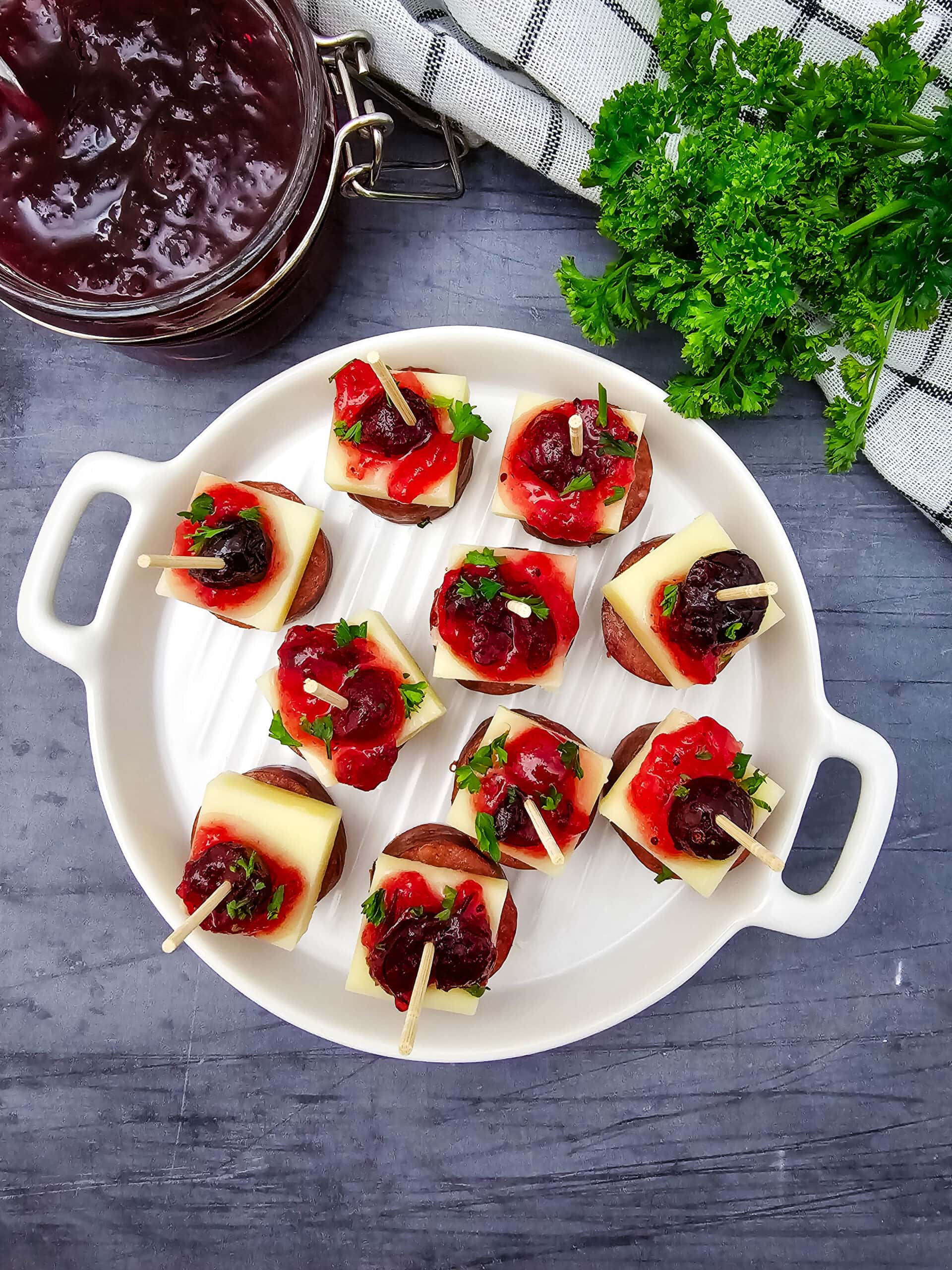 An overhead shot of a white plate that holds skewered appetizers made of sausage slices, cheese cubes, cranberry sauce, and fresh parsley. A jar of sauce and fresh parsley are beside the plate on a gray countertop and checkered cloth.