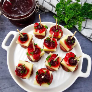 A white plate holds skewers with sausage, cheese, cranberry sauce, and herbs. A jar of cranberry sauce, a bundle of fresh parsley, and a checkered towel are in the background on a dark gray wooden surface.