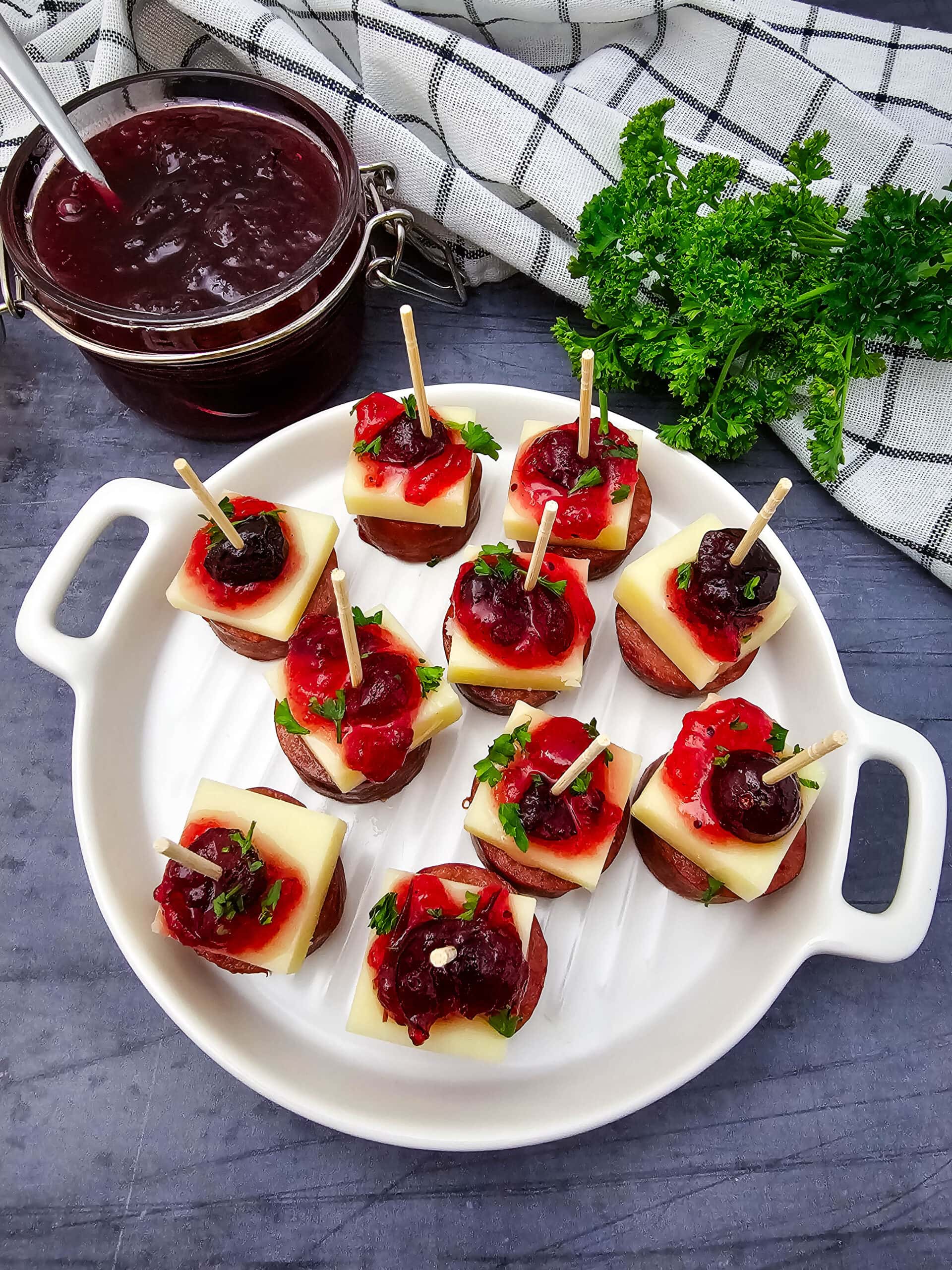 A white plate holds skewers with sausage, cheese, cranberry sauce, and herbs. A jar of cranberry sauce, a bundle of fresh parsley, and a checkered towel are in the background on a dark gray wooden surface.