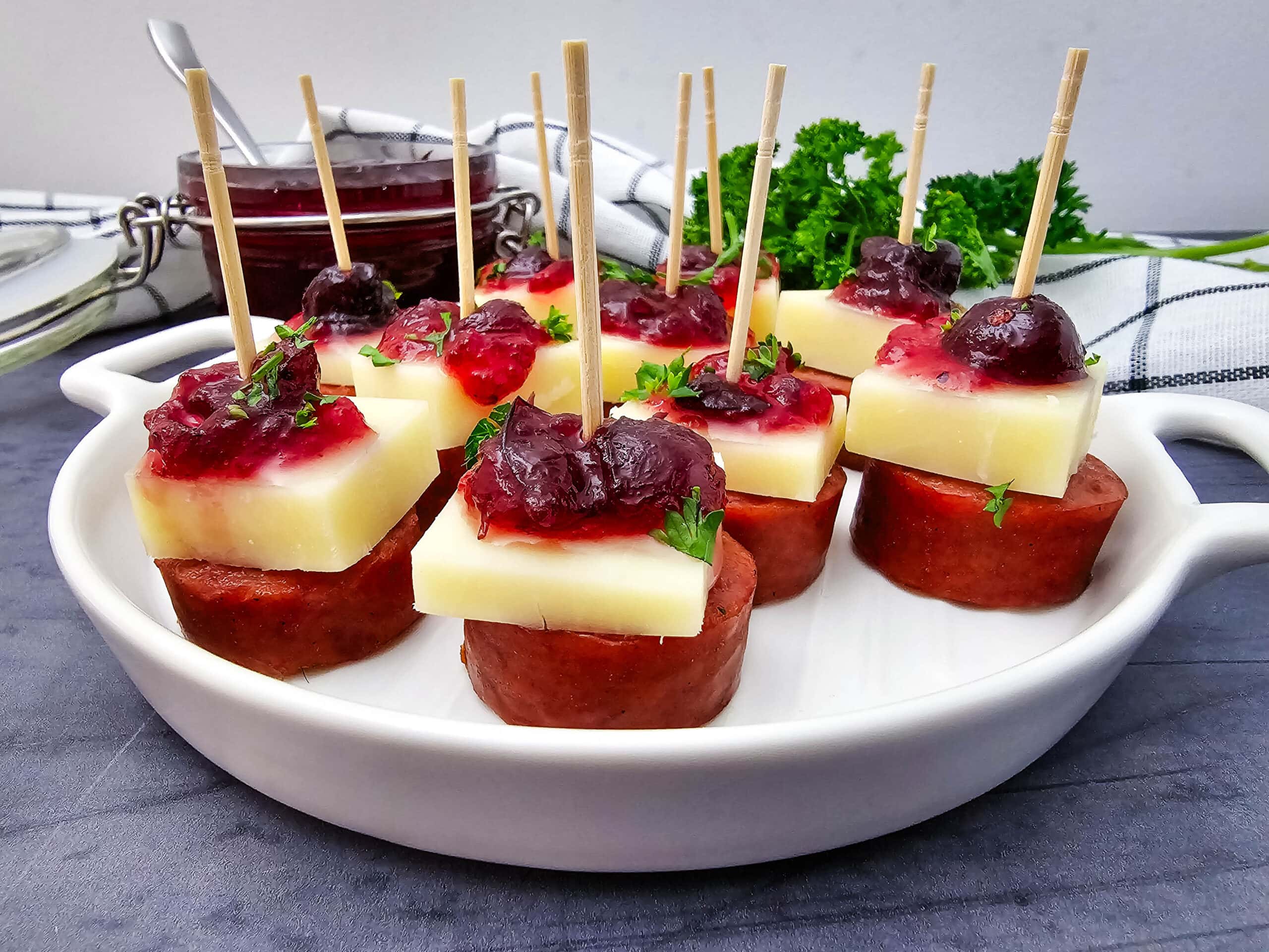 A white dish holds appetizer skewers with sausage slices, cheese cubes, and a dollop of cranberry sauce, garnished with herbs. A jar of cranberry sauce and some parsley are in the background on top of a striped cloth.