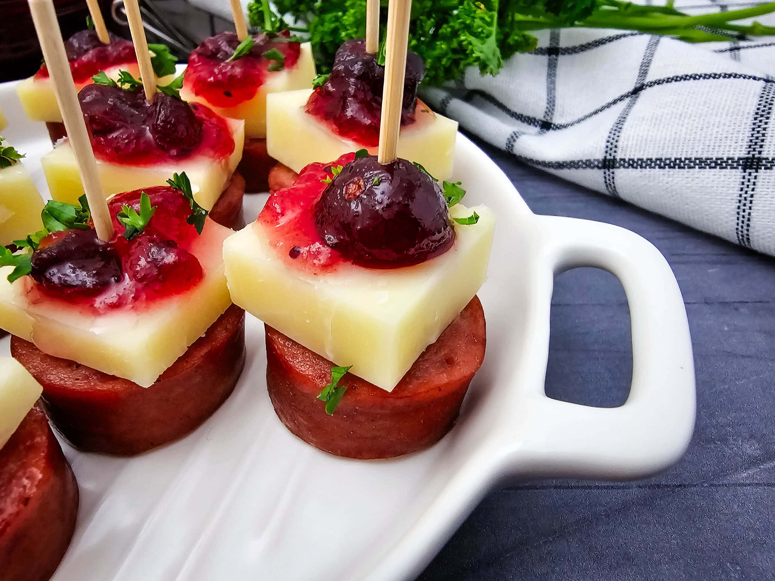 A close up of appetizers on a white tray: skewers with sausage slices, topped with cubes of cheese, cranberry sauce, and garnished with chopped parsley. A checkered cloth and greenery are in the background.