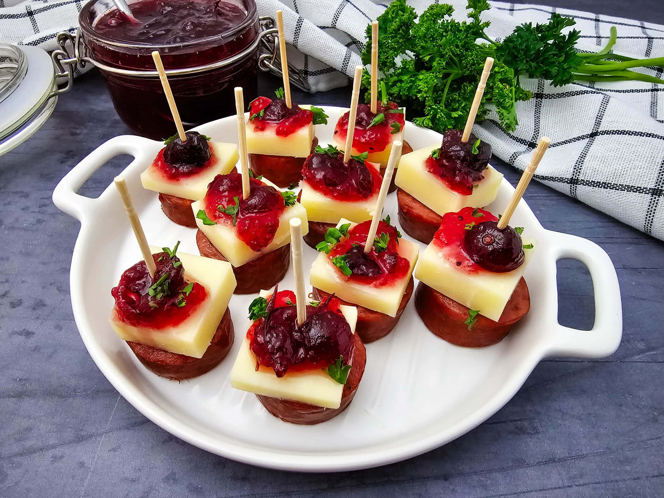 A white plate holds appetizers made of sausage slices, cheese cubes, and a dollop of cranberry sauce, each skewered with a toothpick. Fresh herbs are sprinkled on top, with a jar of cranberry sauce and fresh parsley in the background on a checkered hand towel.