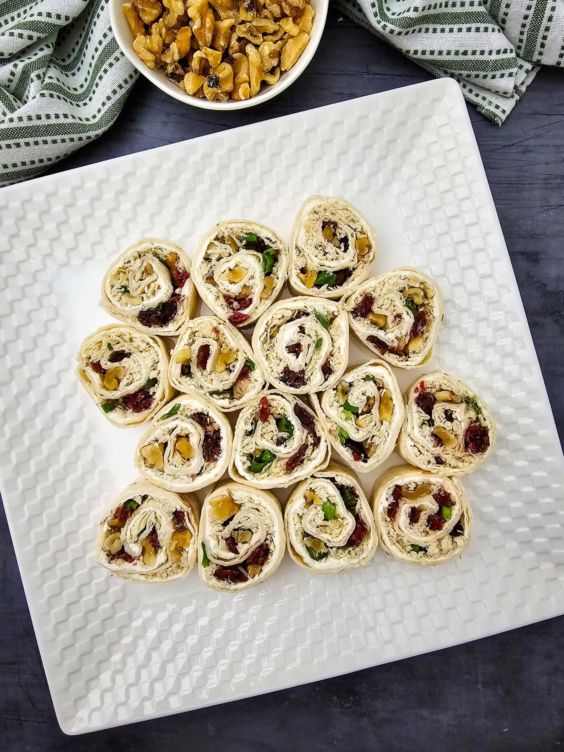 Overhead shot of a white plate displays 15 pinwheel appetizers filled with greens, dried cranberries, and chopped nuts. A bowl of walnuts and a green-striped towel are in the background on a dark surface.