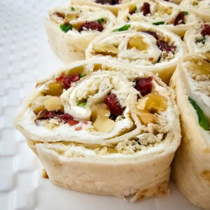 Close-up of sliced pinwheel appetizers on a white plate, made with tortillas, cream cheese, shredded chicken, dried cranberries, chopped green onions, and nuts.