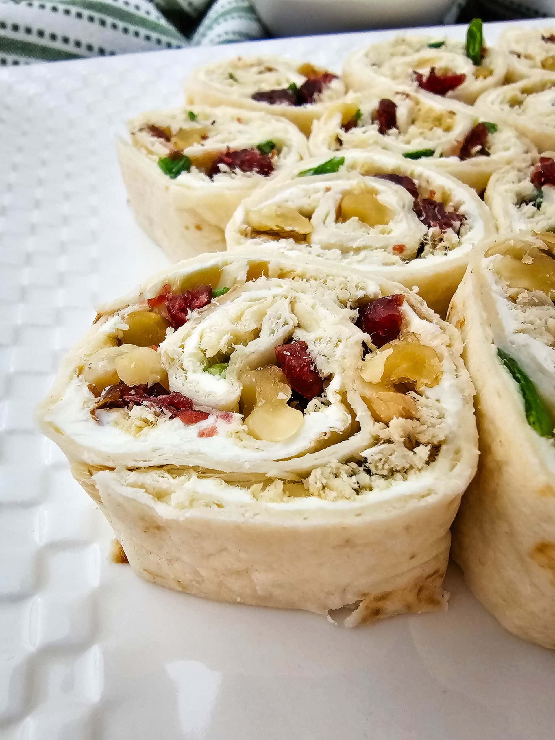 Close-up of sliced pinwheel appetizers on a white plate, made with tortillas, cream cheese, shredded chicken, dried cranberries, chopped green onions, and nuts.