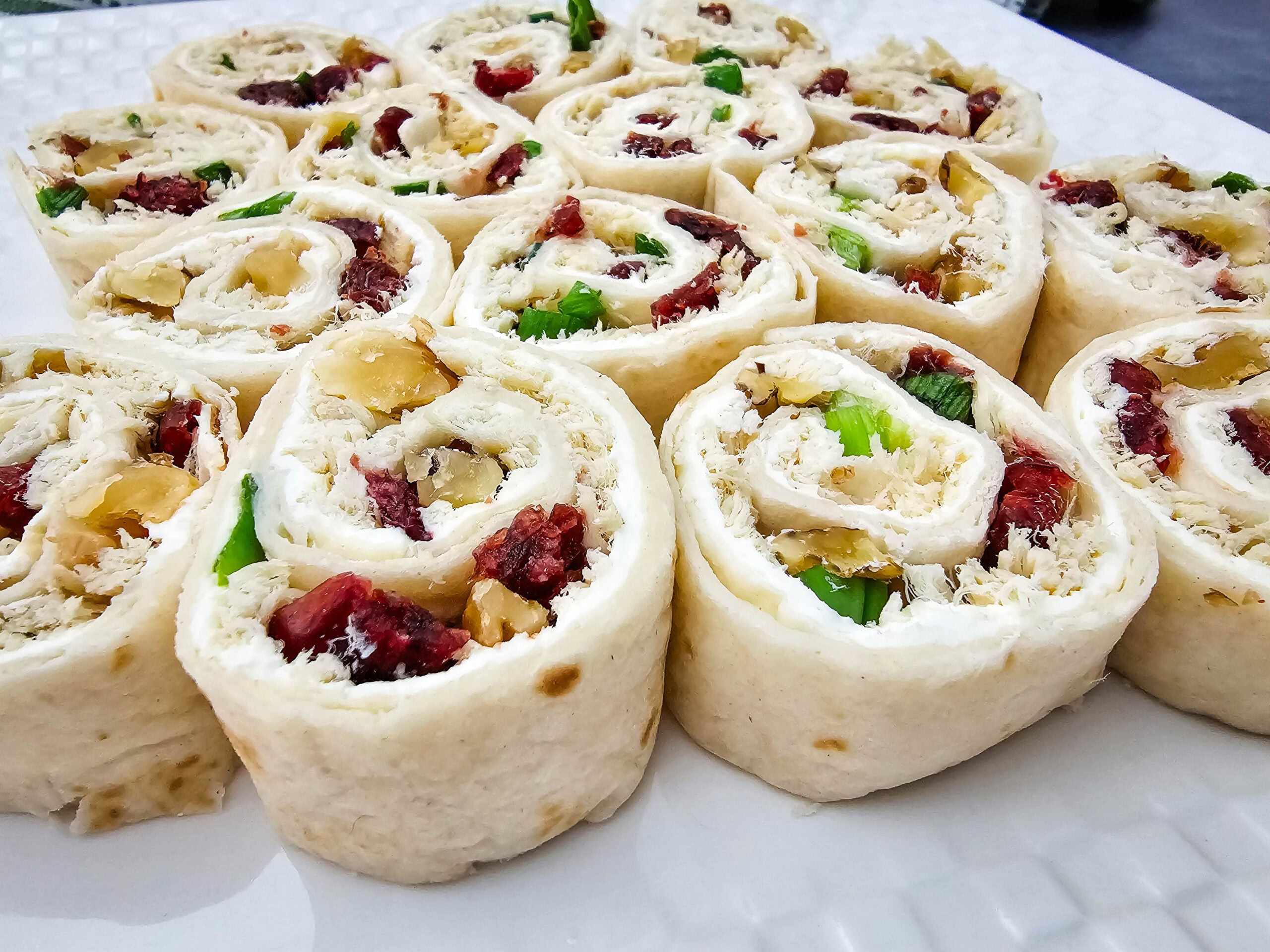 A close-up of several tortilla pinwheels filled with shredded chicken, dried cranberries, green onions, and cream cheese, arranged neatly on a white plate.