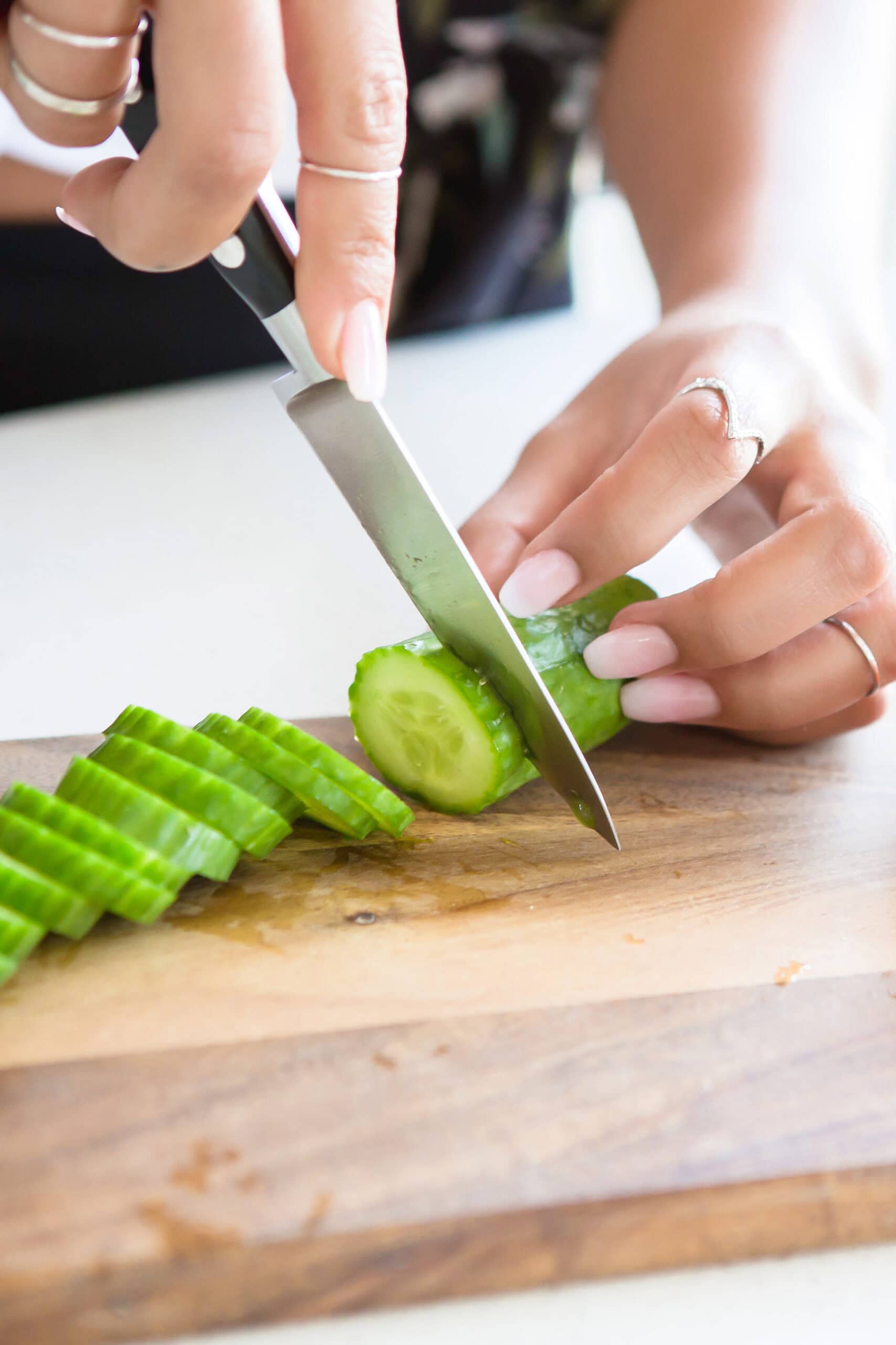 A person with manicured nails slices a cucumber on a wooden cutting board using a knife. Several cucumber slices are already cut and lying on the board.