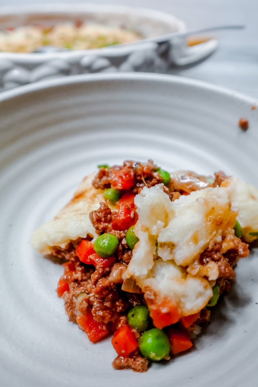 A single serving of shepherds pie on a white plate, featuring ground meat, peas, and diced carrots, topped with creamy mashed potatoes. The casserole dish is blurred in the background.