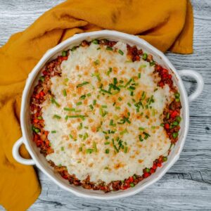 A round casserole dish filled with shepherds pie, topped with mashed potatoes, melted golden cheese and chopped chives, sits on a wooden surface next to a folded mustard-yellow cloth napkin.