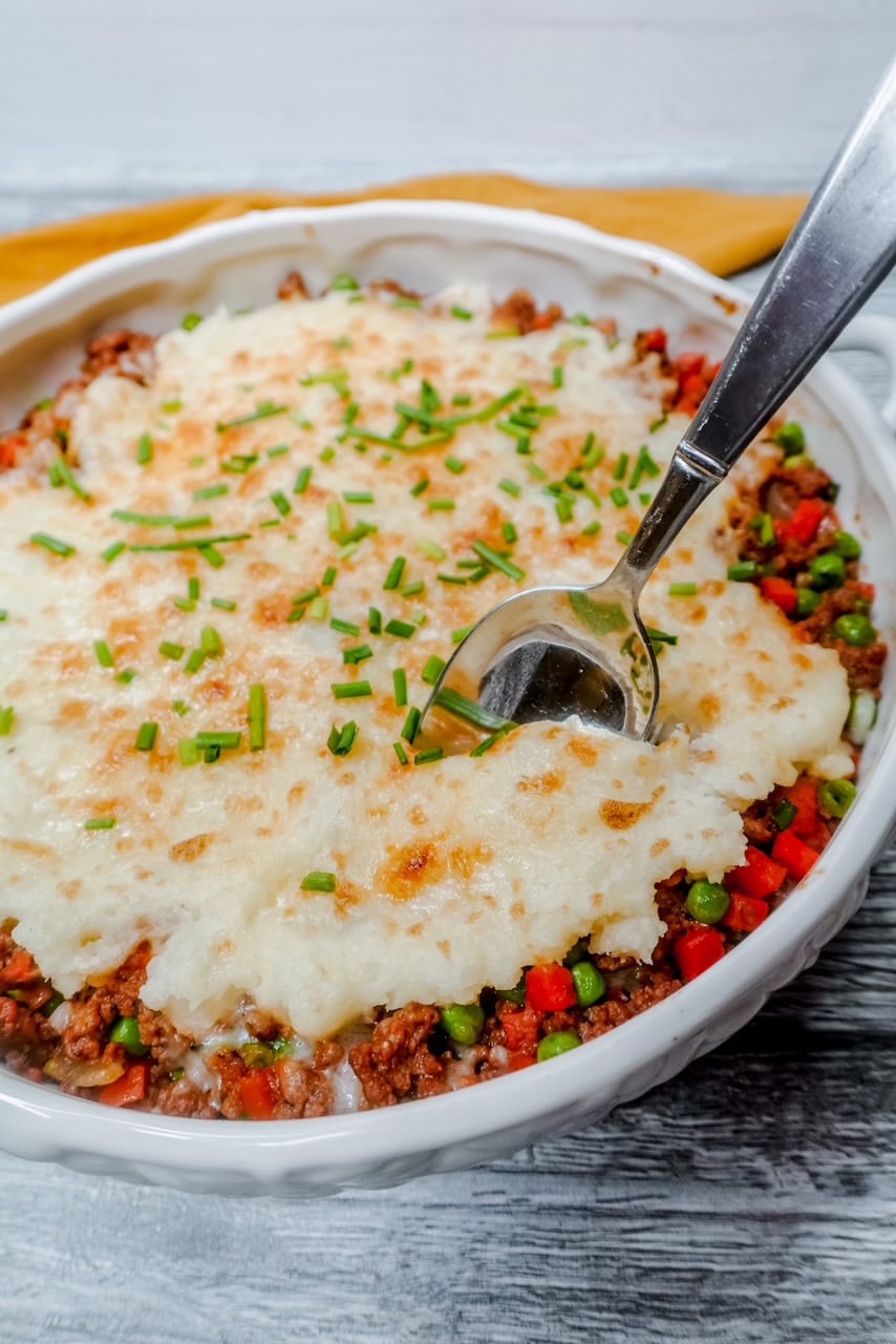 A white dish filled with shepherd&rsquo;s pie, topped with creamy mashed potatoes and chopped chives, showing a spoon scooping into the ground beef and mixed vegetables underneath.