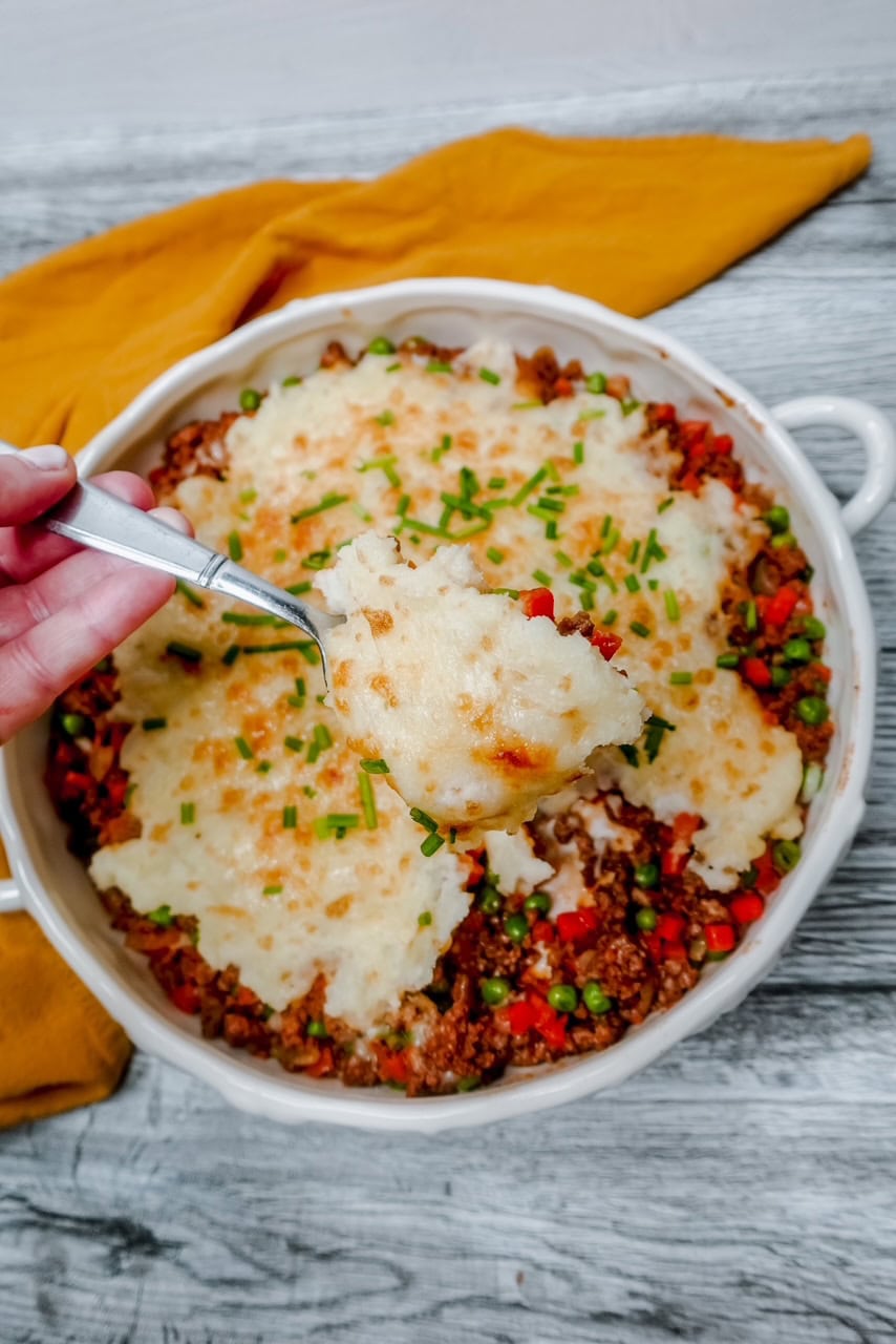 A hand holds a spoonful of shepherd’s pie above a round baking dish filled with the casserole, topped with mashed potatoes, melted cheese, and chopped chives. A yellow cloth lies next to casserole dish on the light gray wooden tabletop.
